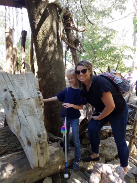 Brittany Clarkson with daughter Addy in a park, Addy touching a tactile wooden sculpture that resembles a dragon