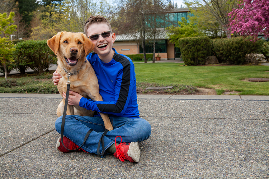 adolescent boy wearing sunglasses sitting on the ground next to a golden retriever dog on a leash