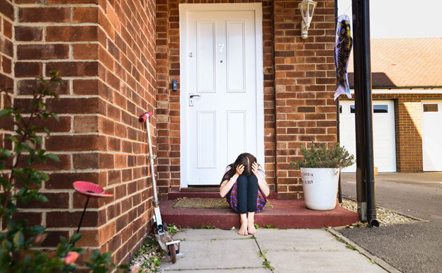 An upset child is sitting on the step in front of their home. The child is holding their head in their hands and resting their forehead on their knees. 