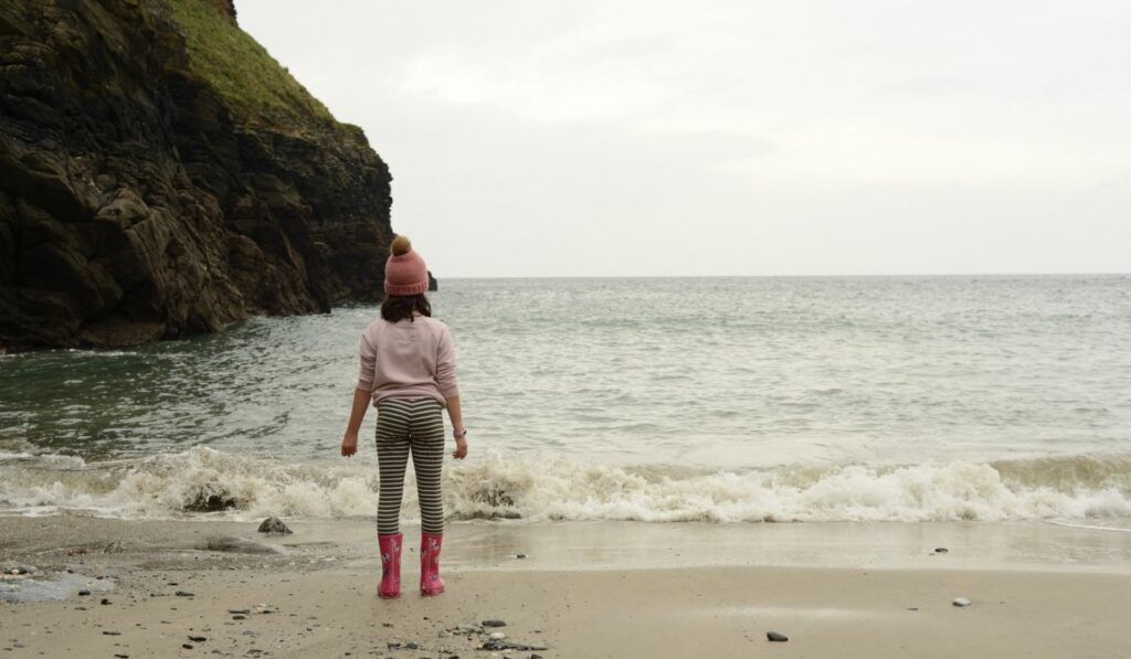 Young child in a knit cap, sweatshirt, and rain boots is standing on the edge of the ocean looking out at the ocean as waves roll up at her feet.
