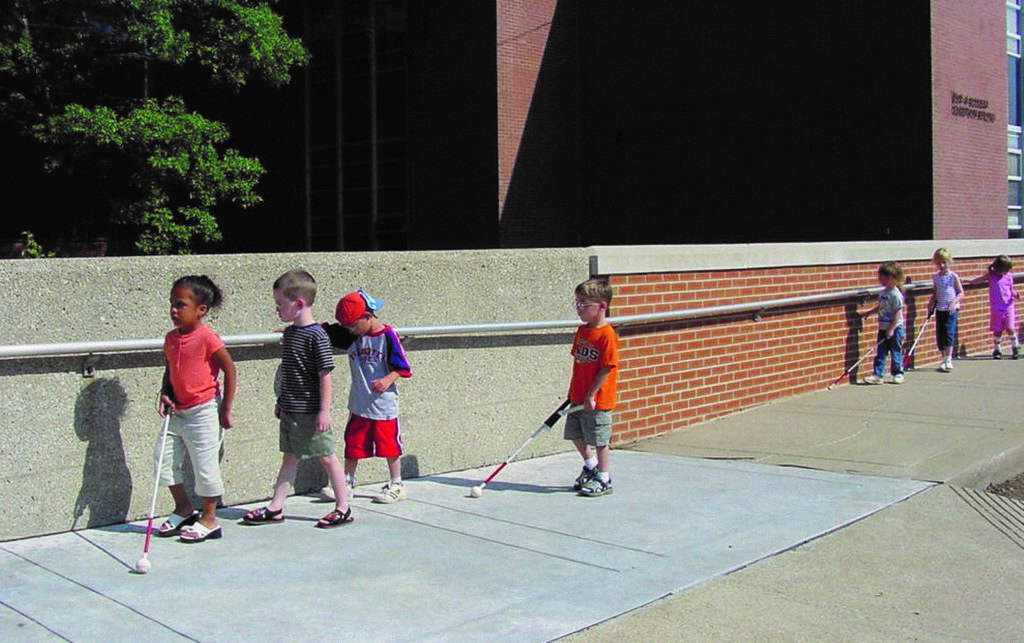 A group of children walking outside along a brick wall using white canes. 