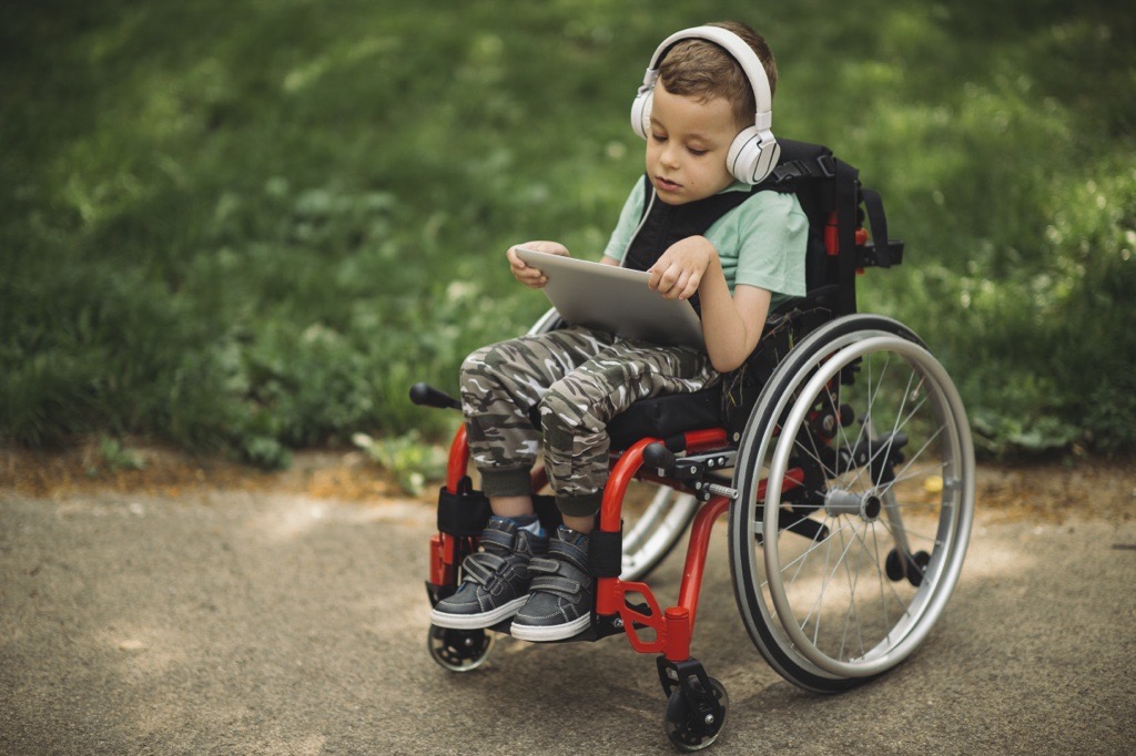 Young child in a wheelchair holding a tablet and wearing headphones.