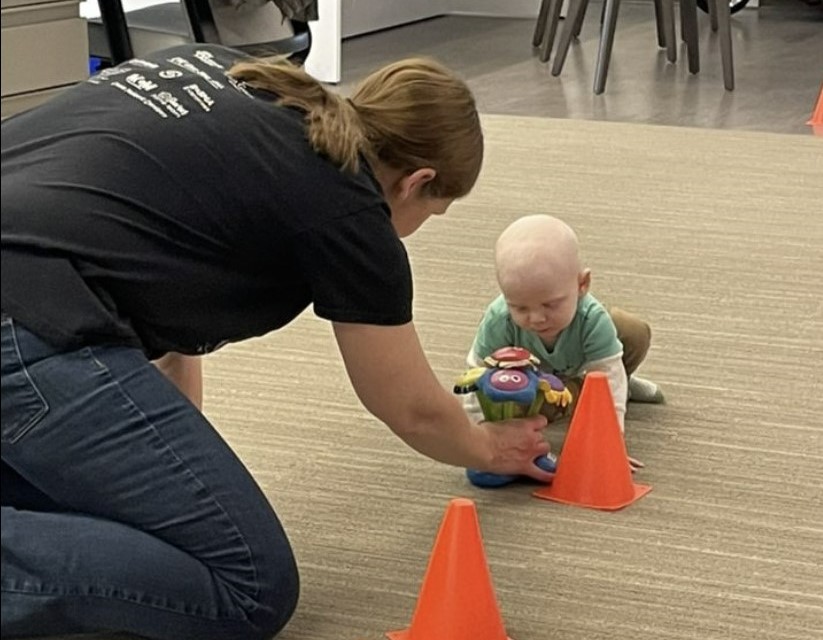 Photo of woman holding a toy with a toddler on the floor in front of an orange cone. 