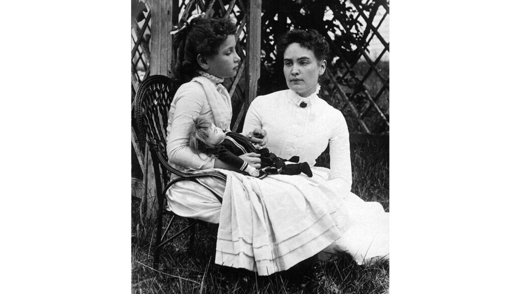 Hellen Keller sitting in a chair holding a doll with her teacher, Anne Sullivan sitting beside her. 