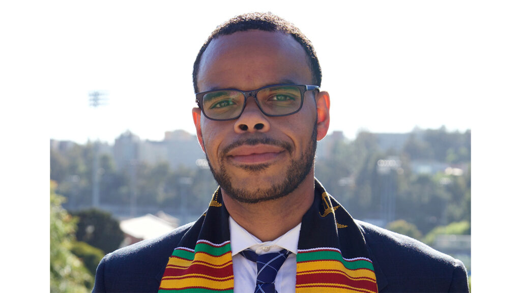 An Afro-Latino man with a short haircut and beard wearing black-framed glasses stands proudly in his graduation gown. A black scarf with multi-colored stripes reads “Luskin Black Caucus at UCLA” on one side and “Master of Social Welfare 2021” on the other. 