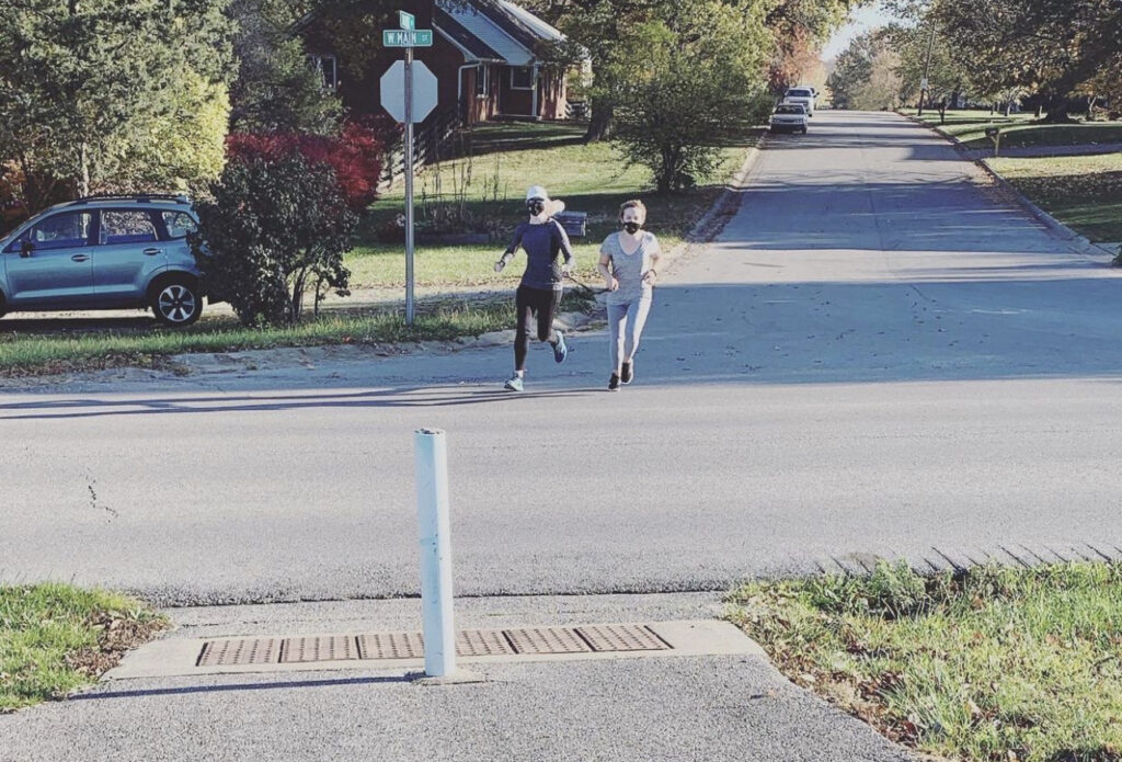 Two girls run towards the camera on a grey road. The girl on the right is in light grey and she is guiding the girl on left with a black tether. The girl on the left has on a white hat, dark sunglasses, and dark grey and black clothing. The background is a neighborhood with green front yards.