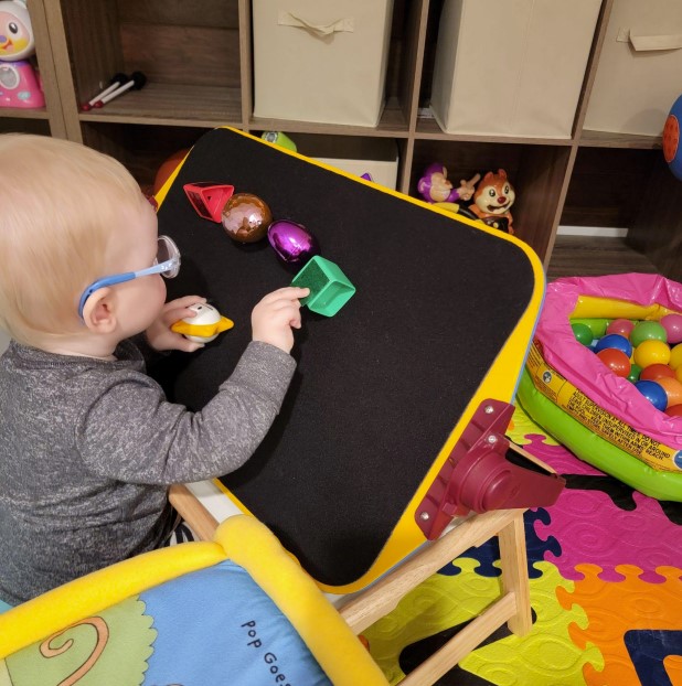 Dominic is sitting on the floor and playing with black felt part of the APH All-In-one board. He is reaching the green square.