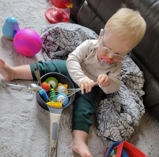 Dominic sitting on the rug and playing with spoons, a pot, and a whisk.