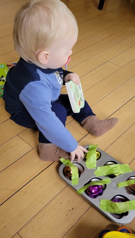 Dominic is playing with a muffin pan. In each spot of the muffin pan, there is an egg shaker taped inside.