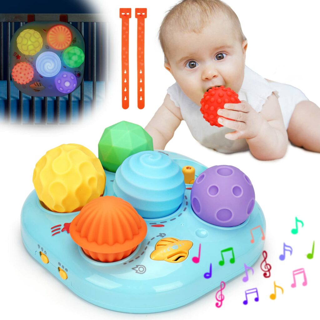 A baby with a textured ball in their mouth laying behind the blue musical toy with 5 different textured balls that light up when pushed.  