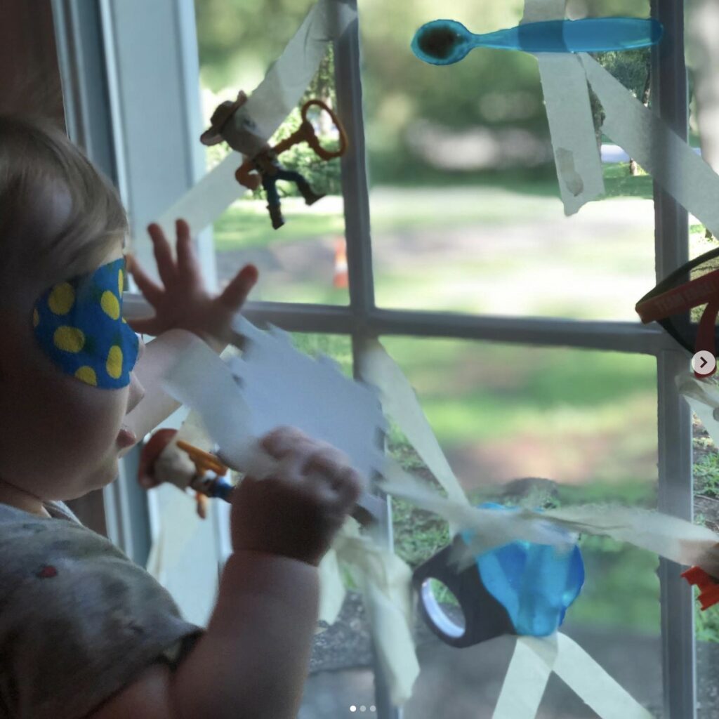 Sutton Sea wearing an eye patch standing against a window where an array of toys are taped to the window for her to locate and pull off.