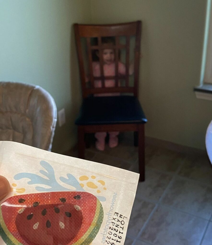 Sutton Sea at 18 months hiding behind a chair (that you can clearly see her through) from her mom whose hand is sneaking into the picture holding an eye patch.  