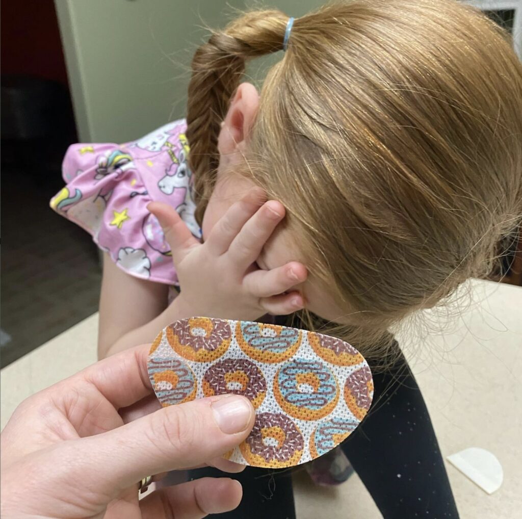 Sutton Sea at 3 years old sitting on a counter covering her eyes as her mom's hand sneaks in the picture holding an eye patch.