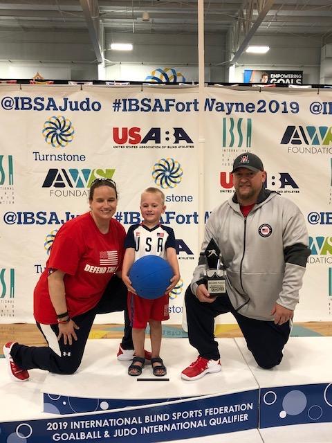 Coach Jake, his wife, and child on a winner’s platform. Text: "2019 International Blind Sports Federation Golball & Judo International Qualifier"