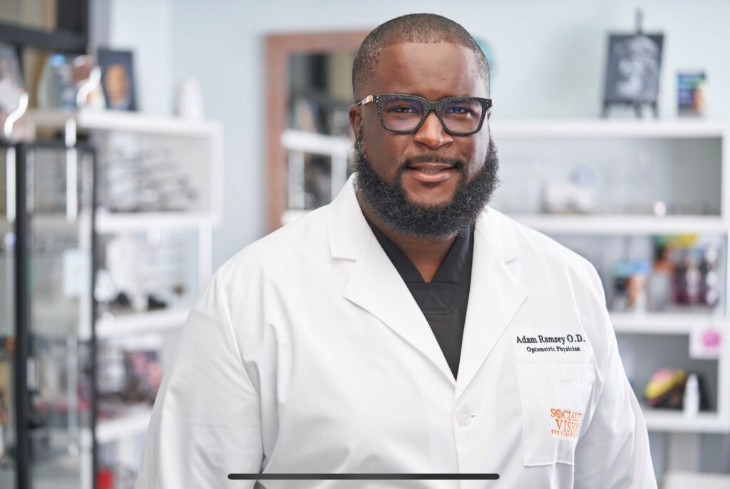 Individual wearing glasses and a white lab coat with embroidered "Adam Ramsey O.D."