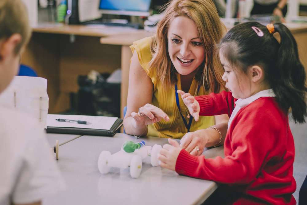 Teacher talks to young student in a red sweater who is touching a balloon-powered model car