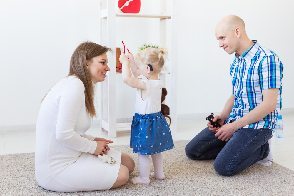 Child with hearing aids playfully gives eyeglasses to an adult 